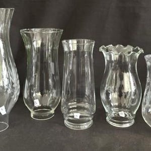 May include: Five clear glass hurricane lamp shades arranged in a row. Each shade features a distinct shape, including flared, straight, and ruffled rims. Some shades have etched floral patterns, and each piece is labeled with a small white numbered tag.