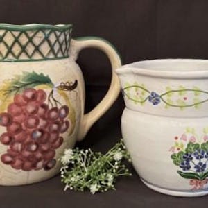 May include: Two ceramic jugs with floral and grape designs. The jug on the left has a green lattice design around the top and a cluster of red grapes on the front. The jug on the right has a white background with a green vine and blue flowers around the top and a bouquet of blue and pink flowers on the front.