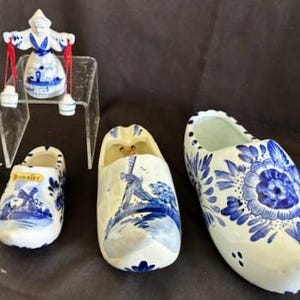 May include: Three blue and white Delftware ceramic clogs, two are taller and one is shorter. The shorter clog has the word "Volendam" written on it. The taller clogs have floral designs and one has a windmill scene.