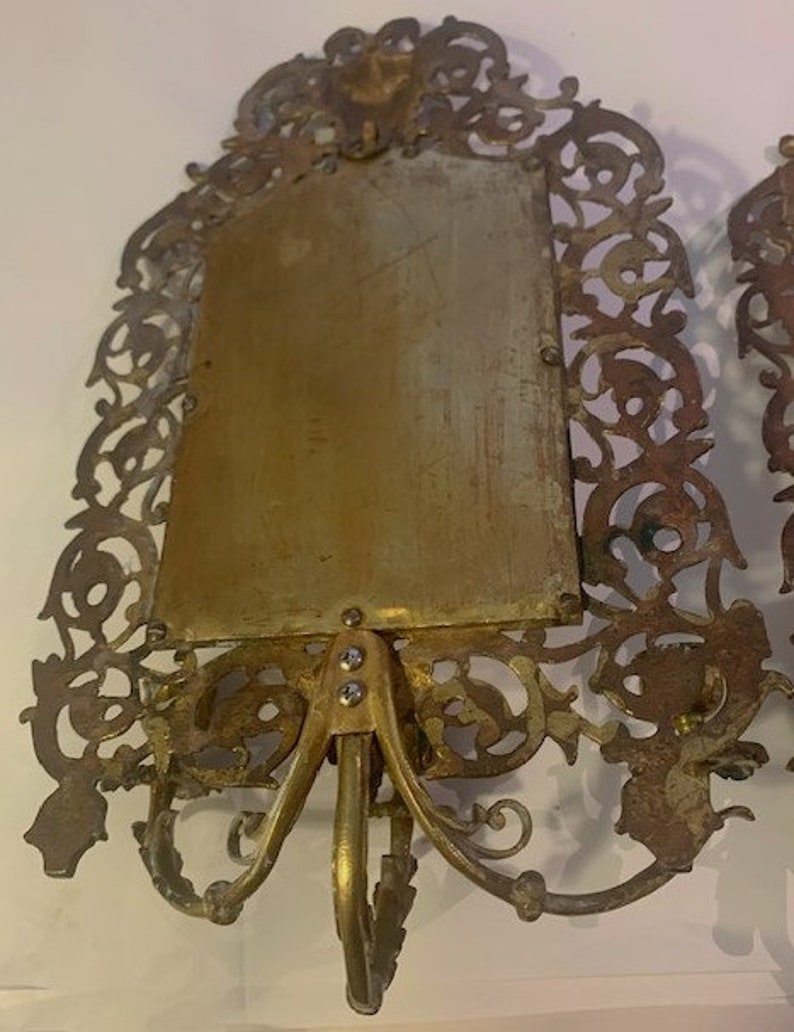 Puede incluir: Un par de marcos de fotos de metal dorado adornados con intrincados dise&ntilde;os de filigrana. Los marcos son rectangulares y est&aacute;n unidos a un soporte decorativo con tres patas.