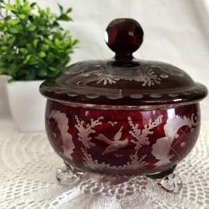 May include: A deep red glass candy dish with a matching lid, featuring etched white floral and bird patterns. The round container rests on three clear glass feet and includes a spherical knob on the lid, showcasing a translucent finish.