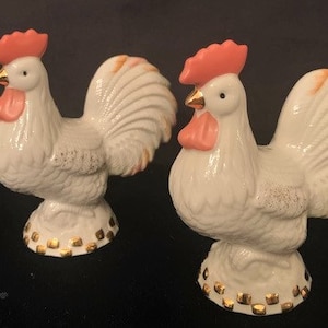 Può includere: Un paio di saliere e pepiere in ceramica bianca a forma di gallo con creste e bargigli rosa. Le basi sono decorate con quadrati dorati.