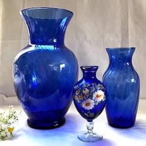 May include: Three blue glass vases of varying sizes. The largest vase is a deep cobalt blue, with a wide, flared opening. The other two vases are a lighter shade of blue. One vase has a painted floral design.