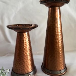 May include: Two copper-colored candle holders with a hammered texture. The holders have a flared, conical shape and are trimmed with a dark metal band at the base and top. The taller holder is approximately 12 inches tall.