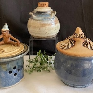 May include: Three ceramic pottery pieces. A blue and white ceramic jar with a lid featuring a small ceramic gnome. A blue and white ceramic mug with a lid featuring a ceramic dragon. A blue and white ceramic jar with a cork lid.