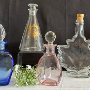May include: Four glass bottles with different shapes and colors. The bottles are clear, blue, pink, and a clear bottle shaped like a maple leaf. The bottles have stoppers and are arranged on a surface.