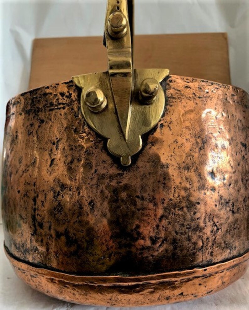May include: A vintage copper pot with a brass handle. The pot has a hammered finish and is showing signs of age.