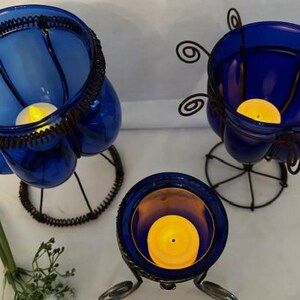 May include: Three blue glass candle holders with wire frames. Each holder has a small, round, white candle inside.