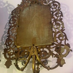 Puede incluir: Un marco de fotos de metal dorado con un dise&ntilde;o ornamentado y filigrana. El marco tiene una abertura rectangular para una foto y una base decorativa con tres patas.