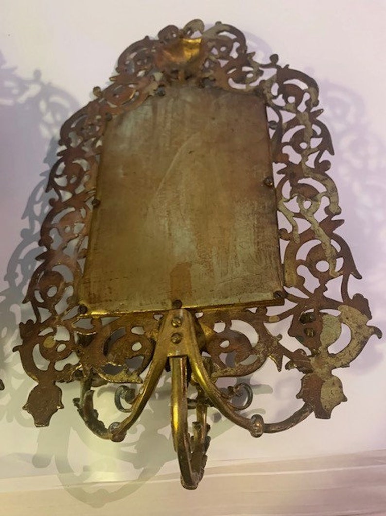 Puede incluir: Un marco de fotos de metal dorado con un dise&ntilde;o ornamentado y filigrana. El marco tiene una abertura rectangular para una foto y una base decorativa con tres patas.