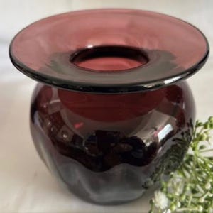 Puede incluir: Un jarr&oacute;n de vidrio de color morado oscuro con una abertura ancha y acampanada y un cuerpo redondeado. El jarr&oacute;n tiene una superficie texturizada y ondulada.