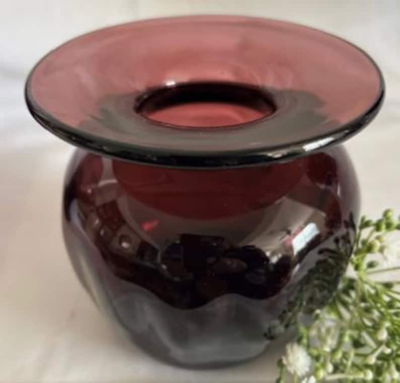 Puede incluir: Un jarr&oacute;n de vidrio de color morado oscuro con una abertura ancha y acampanada y un cuerpo redondeado. El jarr&oacute;n tiene una superficie texturizada y ondulada.