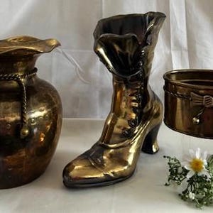 May include: A collection of antique brass decorative items. Includes a vase with a ruffled rim and rope detail, a boot-shaped planter, and a small container with a bow accent. A small floral arrangement is also present.