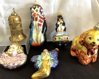 May include: A collection of six glass Christmas ornaments including a gold bell, a penguin with a red scarf, a penguin on a globe, a dog with a red collar, a fairy with blue wings, and a figure with a ruffled collar, all featuring metallic finishes.