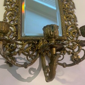 Puede incluir: Un espejo de pared dorado con adornos de volutas y tres candelabros. El espejo es rectangular y tiene un borde biselado.