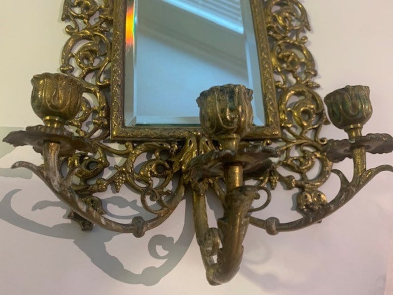 Puede incluir: Un espejo de pared dorado con adornos de volutas y tres candelabros. El espejo es rectangular y tiene un borde biselado.