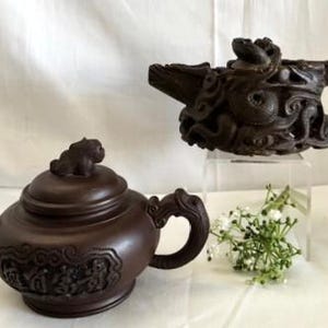 May include: Two brown ceramic teapots with intricate designs. One teapot features a dragon design, while the other has a more traditional design with a small animal on the lid.