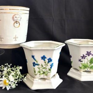 May include: Three white ceramic planters. One cylindrical planter with gold trim, a lion's head ring handle, and fleur-de-lis designs. Two hexagonal planters with floral designs, one with blue flowers and the other with purple flowers, on white pedestals.