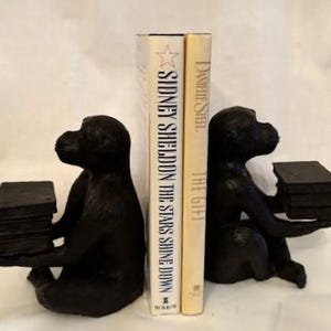May include: A pair of black monkey bookends holding books. The book on the left is "Sidney Sheldon The Stars Shine Down" and the book on the right is "Danielle Steel The Gift".