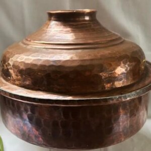 May include: A hammered copper cooking pot with a lid and two ornate handles. The pot has a rounded base and a flared rim. The lid has a rounded top and a small, cylindrical handle. The handles are gold-colored.