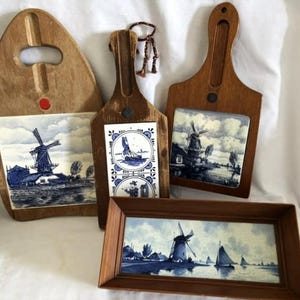 May include: A collection of wooden cutting boards and a framed picture, each featuring blue and white Delftware-style windmill scenes. The boards have handles and decorative tiles. The framed picture is rectangular. The items are arranged on a white surface.