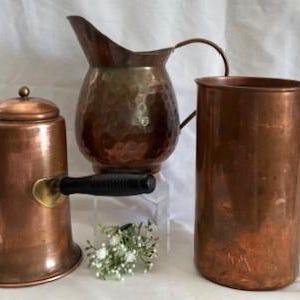 May include: Three copper kitchenware items: a teapot with a black handle, a hammered pitcher, and a tall mug with a handle. The items are displayed on a white surface. The teapot has a spout and a lid with a small knob.