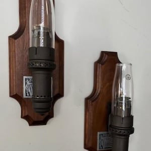 May include: Two antique wall sconces. Each sconce features a dark brown wooden backing with a decorative shape. A dark metal lamp fixture is attached, topped with a clear glass chimney. A small metal plate is affixed to the wooden backing.