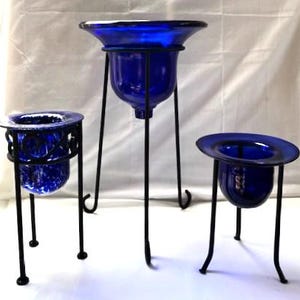 May include: Three cobalt blue glass planters with black metal stands. The planters vary in size and shape, with a flared rim and a rounded base. The stands have a simple, elegant design.