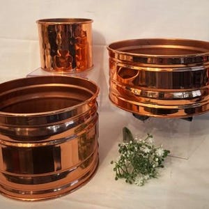 Puede incluir: Tres macetas de cobre de diferentes tamaños. La maceta más grande tiene un borde liso y redondeado y una textura martillada. Las dos macetas más pequeñas tienen un borde liso y redondeado y una textura acanalada.