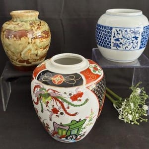 Puede incluir: Tres jarrones de cerámica con diferentes diseños. El jarrón de la izquierda es marrón y verde con un esmalte moteado. El jarrón del centro es blanco con diseños florales rojos, dorados y negros. El jarrón de la derecha es blanco con diseños geométricos azules y dorados.