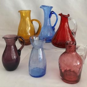 May include: A collection of colorful glass pitchers in various shades of amber, blue, red, and purple. Each pitcher has a handle and a spout for pouring. The pitchers are of different sizes and shapes, suitable for serving beverages.