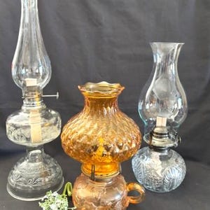 Könnte beinhalten: Drei Öllampen aus Glas in verschiedenen Größen und Designs. Eine ist klar mit einem hohen Schornstein, eine andere ist bernsteinfarben mit rundem Körper, und die dritte ist klar mit einem floralen Muster. Ein kleiner Zweig weißer Blumen liegt davor.