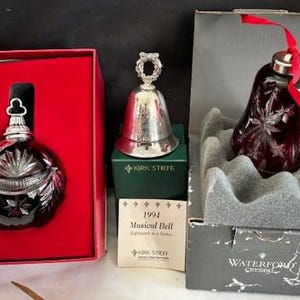 Puede incluir: Dos adornos navideños en sus cajas originales. Uno es una campana de cristal roja con un diseño de copo de nieve y el texto "Waterford Crystal". El otro es una campana de plata con el texto "Kirk Stieff 1994 Musical Bell Fifth in a Series".