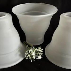 May include: Three frosted glass lamp shades with a bell shape. The shades are white and have a smooth, matte finish. A small bouquet of white flowers rests near the shades. The background is black.