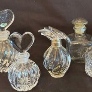 May include: A collection of five clear glass perfume bottles with various stoppers. Two bottles feature heart-shaped stoppers, one has a fan-shaped stopper, one is square with a round stopper, and one is cylindrical with a faceted rectangular stopper.