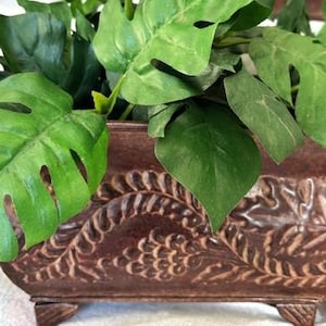 May include: A rectangular, brown metal planter with a detailed embossed design, filled with vibrant green artificial leaves. The planter has small feet and a decorative border. The leaves have a natural appearance, adding a touch of nature to the decor.