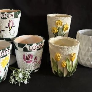 May include: A collection of ceramic flower pots with various designs. Some pots feature painted flowers, the words "HOPE", "FAITH", and "LOVE", and a textured, stone-like pattern. One pot is plain white with a geometric design.