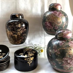 May include: A collection of decorative ceramic jars and dishes. The jars are black with gold floral and landscape designs, and the other jars are dark green with pink floral patterns. The dishes are black with gold floral designs.