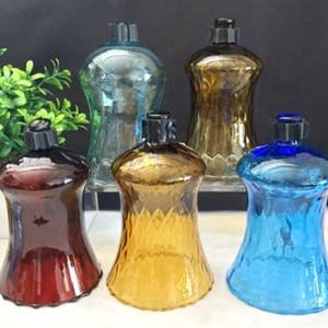 May include: Five glass candle holders in various colors including dark red, amber, bright blue, light blue, and brown. Each holder features a flared shape with a textured diamond pattern on the lower half and a black circular rim at the top opening.