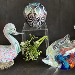 Può includere: Tre figurine di vetro: un cigno bianco e rosa, un delfino di vetro trasparente con vetro colorato all'interno e una sfera di vetro iridescente rotonda con un motivo a spirale.