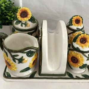May include: A ceramic kitchen set on a rectangular tray featuring a creamer, a napkin holder, and two lidded containers. Each piece is white with hand-painted yellow sunflowers, green leaves, and dark green vine accents along the edges.