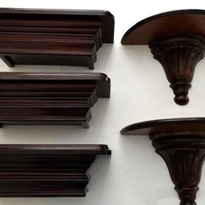 Op de afbeelding: Een verzameling donkerbruine houten planken. Drie zijn rechthoekig met gelaagde lijstwerk, en twee zijn halfrond met sierlijke, gesneden steunen. De planken zijn aan een witte muur bevestigd.