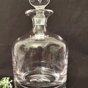 May include: A clear glass decanter with a round stopper. The decanter has a wide base and a narrow neck, with a spherical stopper. The glass is transparent, allowing the contents to be visible. The decanter is likely used for serving beverages.