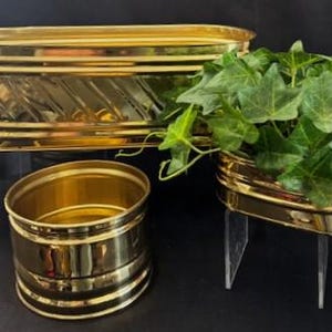 Puede incluir: Tres macetas de metal de color dorado de diferentes tamaños. La maceta más grande es de forma ovalada y tiene un patrón decorativo. La maceta más pequeña es redonda y la maceta del medio tiene forma de corazón. Las tres macetas están sobre soportes de acrílico transparente.