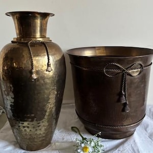 Peut inclure: Deux pièces décoratives antiques en métal de couleur bronze. Un grand vase avec une texture martelée et des détails de corde avec des glands. Une jardinière assortie avec des accents de corde et de glands. Un petit bouquet de fleurs blanches à cœur jaune repose devant.