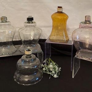 Puede incluir: Una colección de lámparas de aceite de vidrio vintage en varias formas y colores. Las lámparas incluyen tonos claros, ámbar y rosa, con tapas de metal y plástico. Algunas lámparas se exhiben sobre soportes acrílicos transparentes, con una ramita de flores blancas en primer plano.