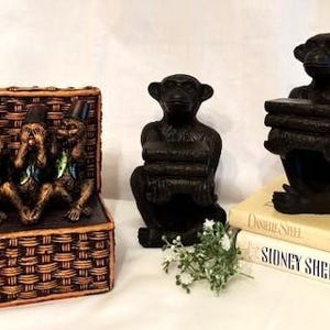 May include: Three small monkey figurines in a woven basket, each with a different hand gesture, representing "see no evil", "hear no evil", and "speak no evil". Two larger black monkey figurines are holding books, acting as bookends.