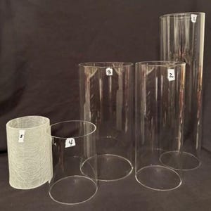 Peut inclure: Ensemble de cinq cylindres en verre transparents de différentes tailles. Quatre sont lisses et transparents, avec des dessus ouverts et des fonds fermés. Un cylindre a une finition texturée et givrée. Les cylindres sont disposés sur une surface sombre.