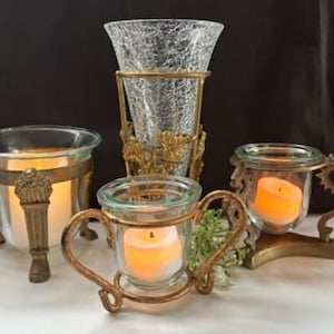 May include: An assortment of decorative candle holders. Includes a tall, textured glass vase in a gold metal stand, and several glass candle holders with ornate gold metal accents. Each holder contains a lit candle.