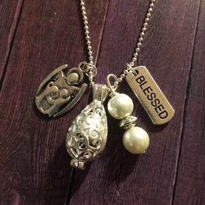 May include: Silver-toned necklace with multiple charms. The charms include an angel, a filigree teardrop-shaped pendant, two faux pearls, and a rectangular tag that reads "BLESSED". The necklace is displayed on a dark wood-grain surface.